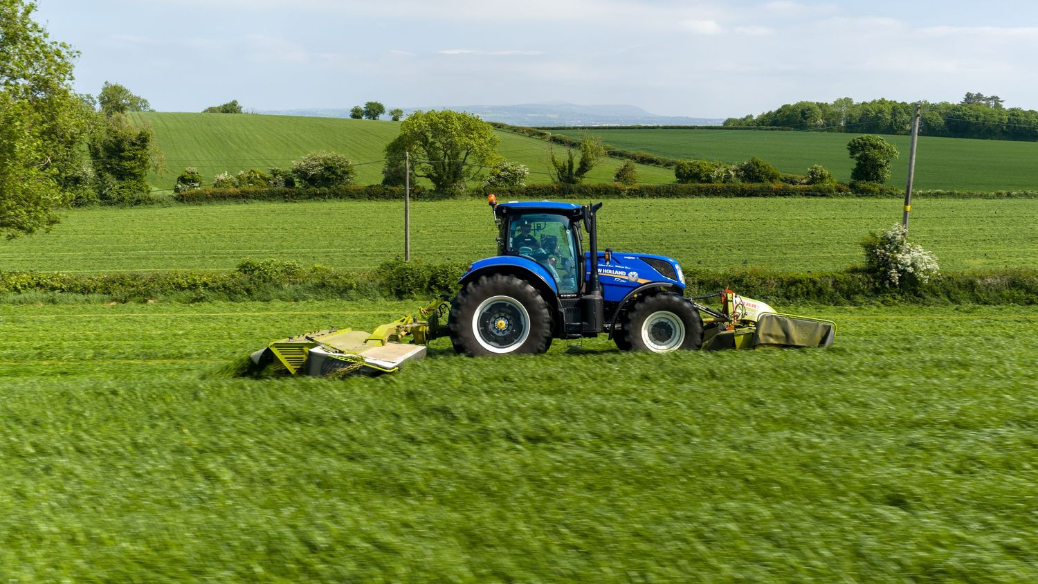 Blue New Holland tractor mowing green fields in rural countryside on an overcast day.