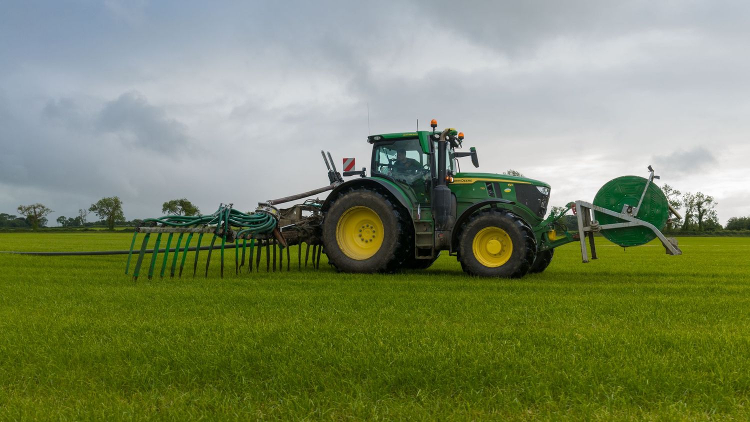 Green John Deere tractor with rake attachment working in a lush field under cloudy skies.