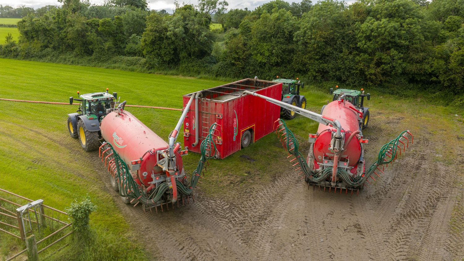 Two red agricultural slurry tankers connected to a red trailer, operated by green tractors in a muddy farm field.