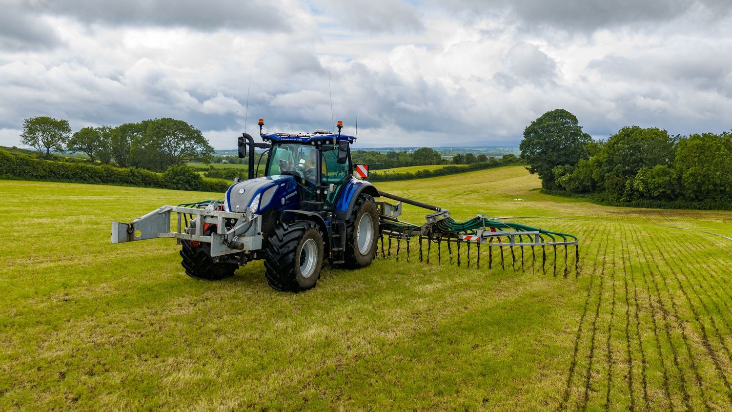 Blue tractor with wide rake attachment working in lush green field under cloudy sky in rural countryside.