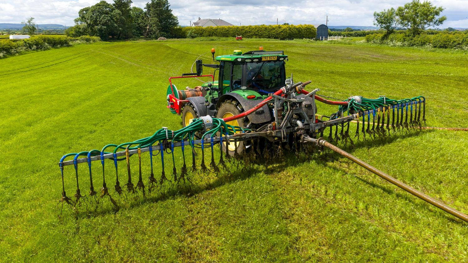Green John Deere tractor with wide rake attachment spreading across lush green field on a sunny day.