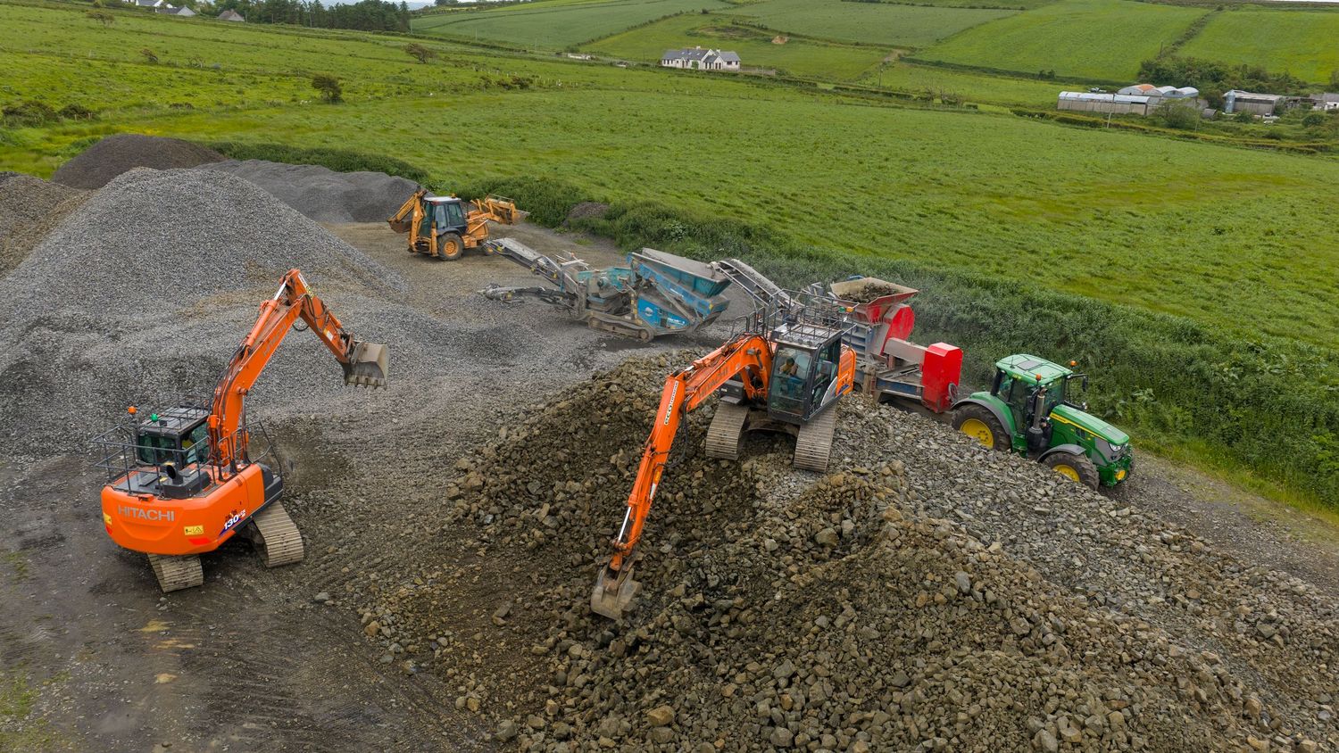 Orange excavators and green tractors moving gravel on a rural construction site surrounded by green fields.