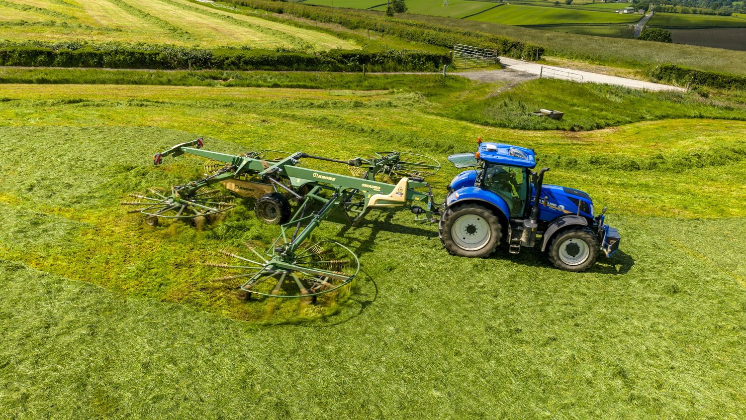 Aerial view of a blue New Holland tractor using a rotary rake to ted grass in a lush green field.