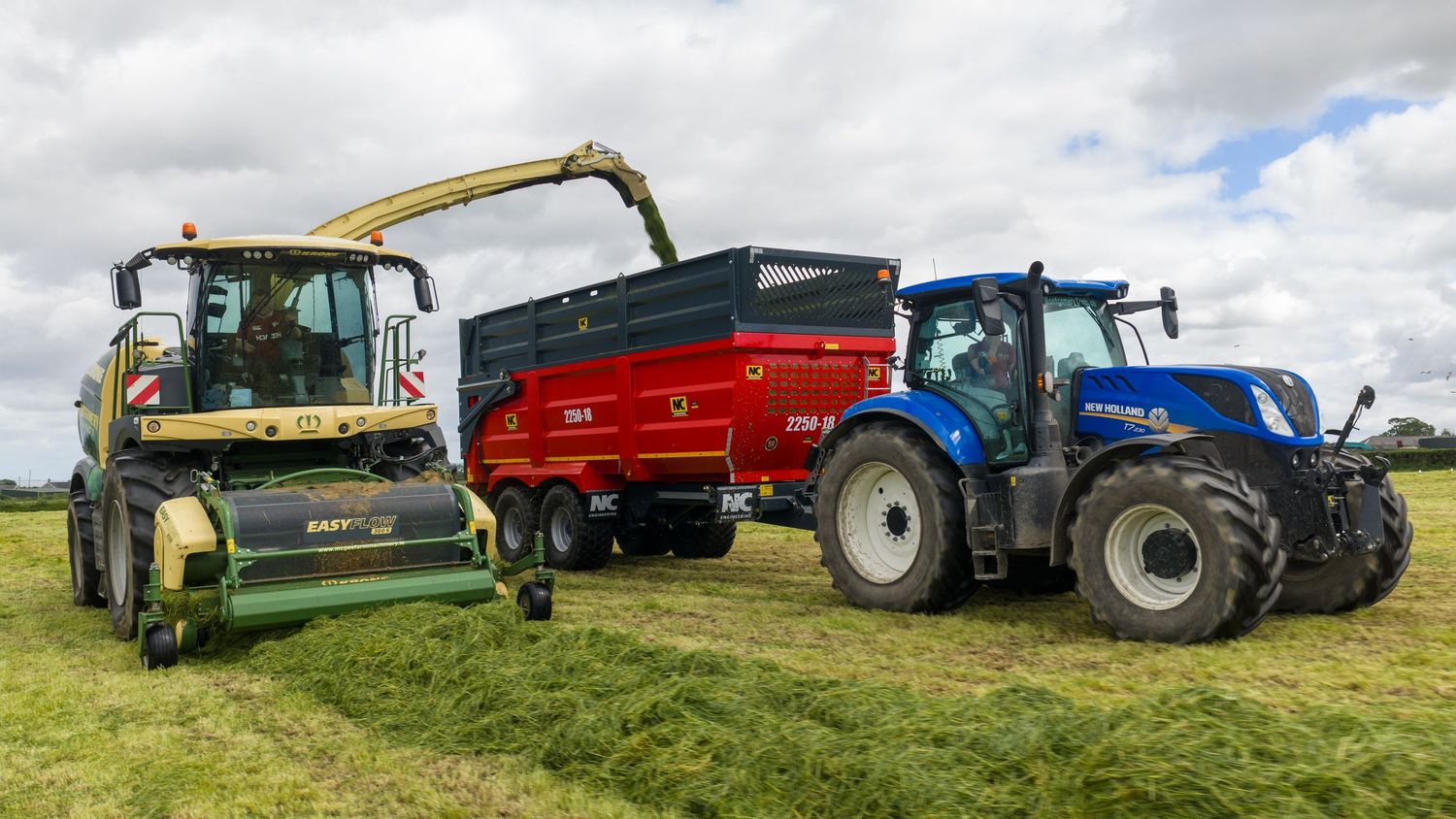 Green and blue tractors harvesting grass silage in a field, with a red trailer collecting cut grass on a sunny day.
