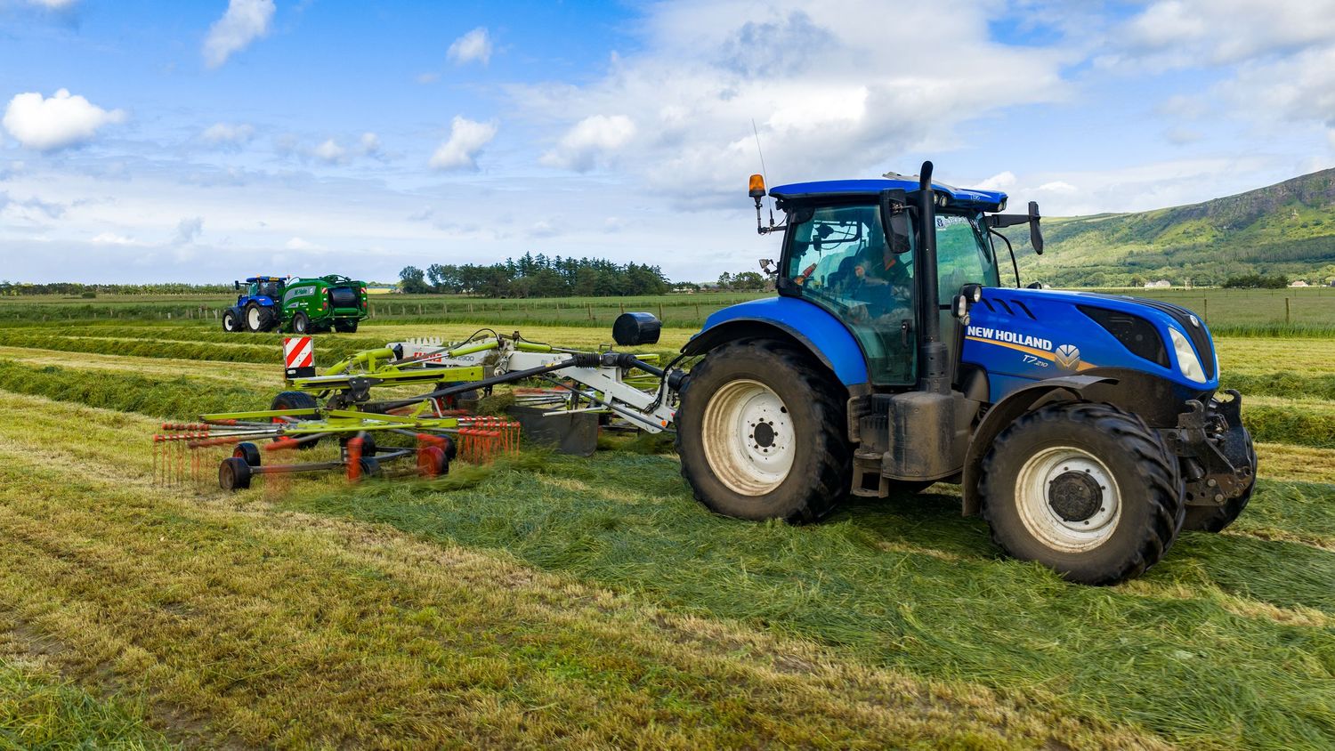Blue New Holland tractor with hay rake attachment working in a green field under a cloudy sky.