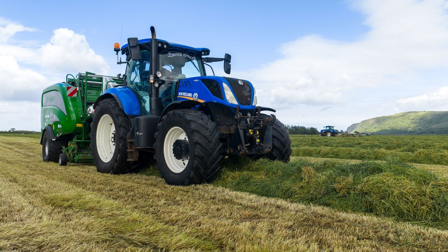Blue New Holland tractor baling hay in a green field on a sunny day with rolling hills in the background.