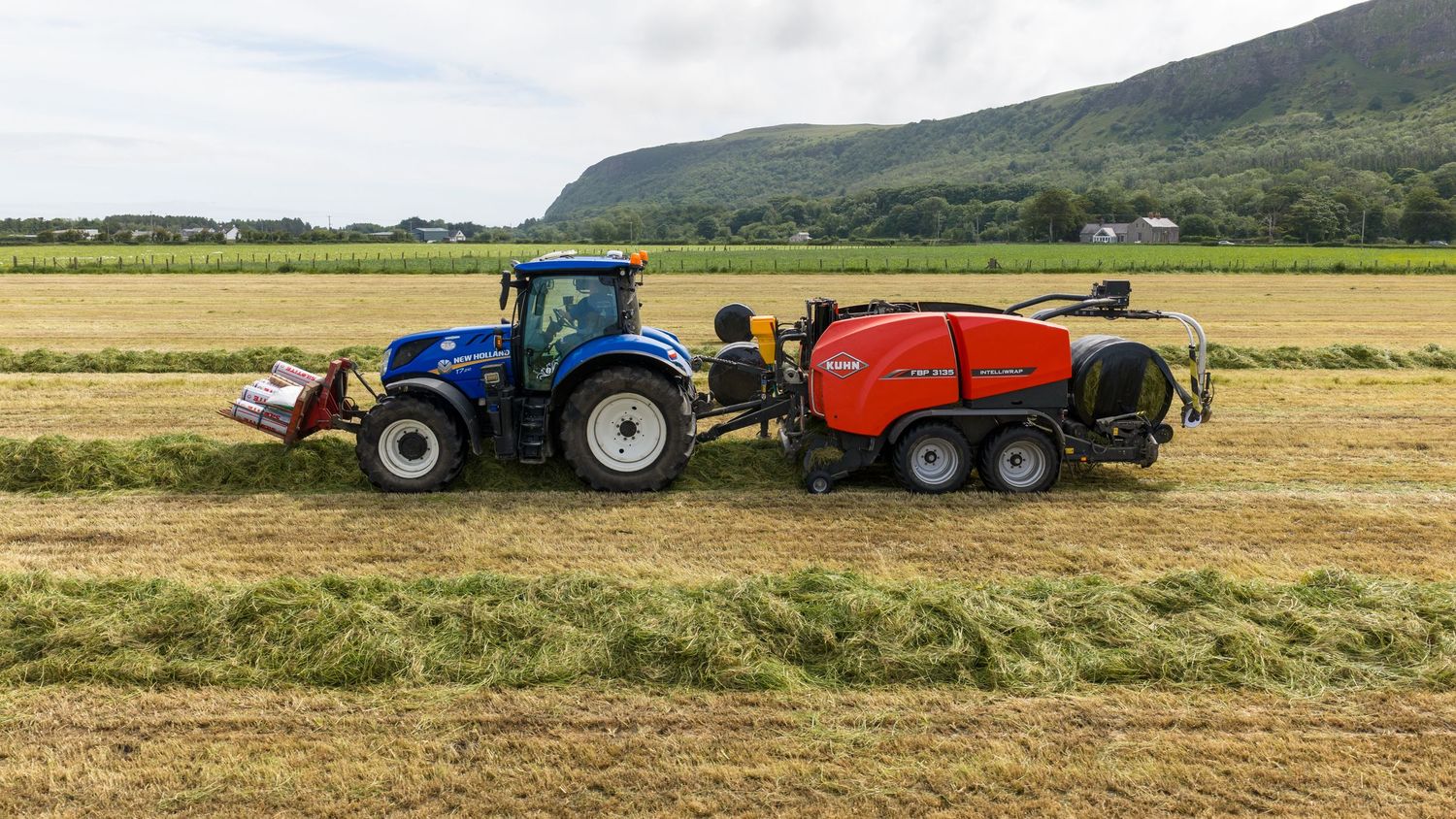 Red and blue New Holland tractor with baler attachment harvesting hay in a green field with mountain backdrop.