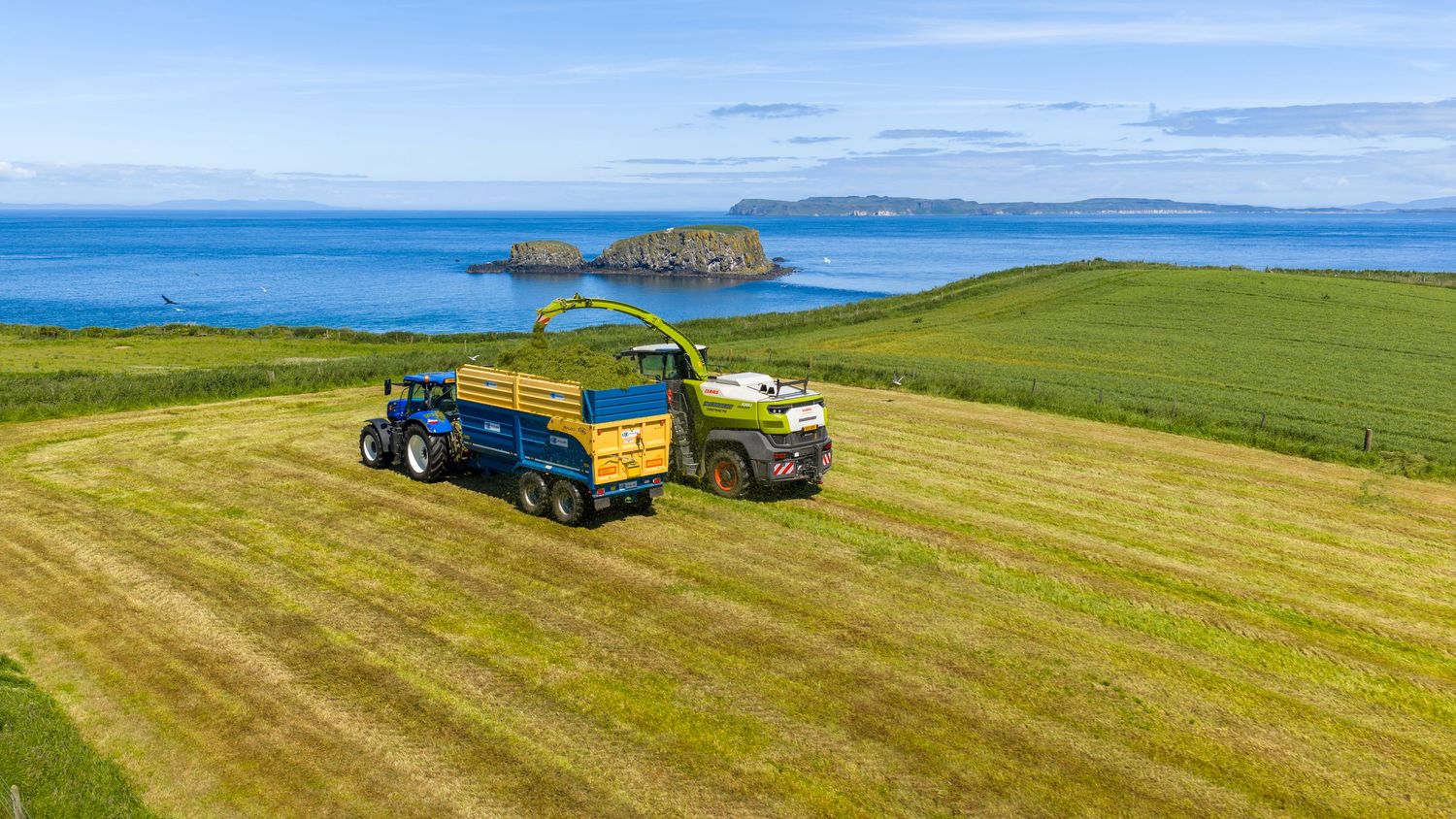 Aerial view of a green forage harvester and tractor working coastal farmland fields with blue ocean and islands beyond.
