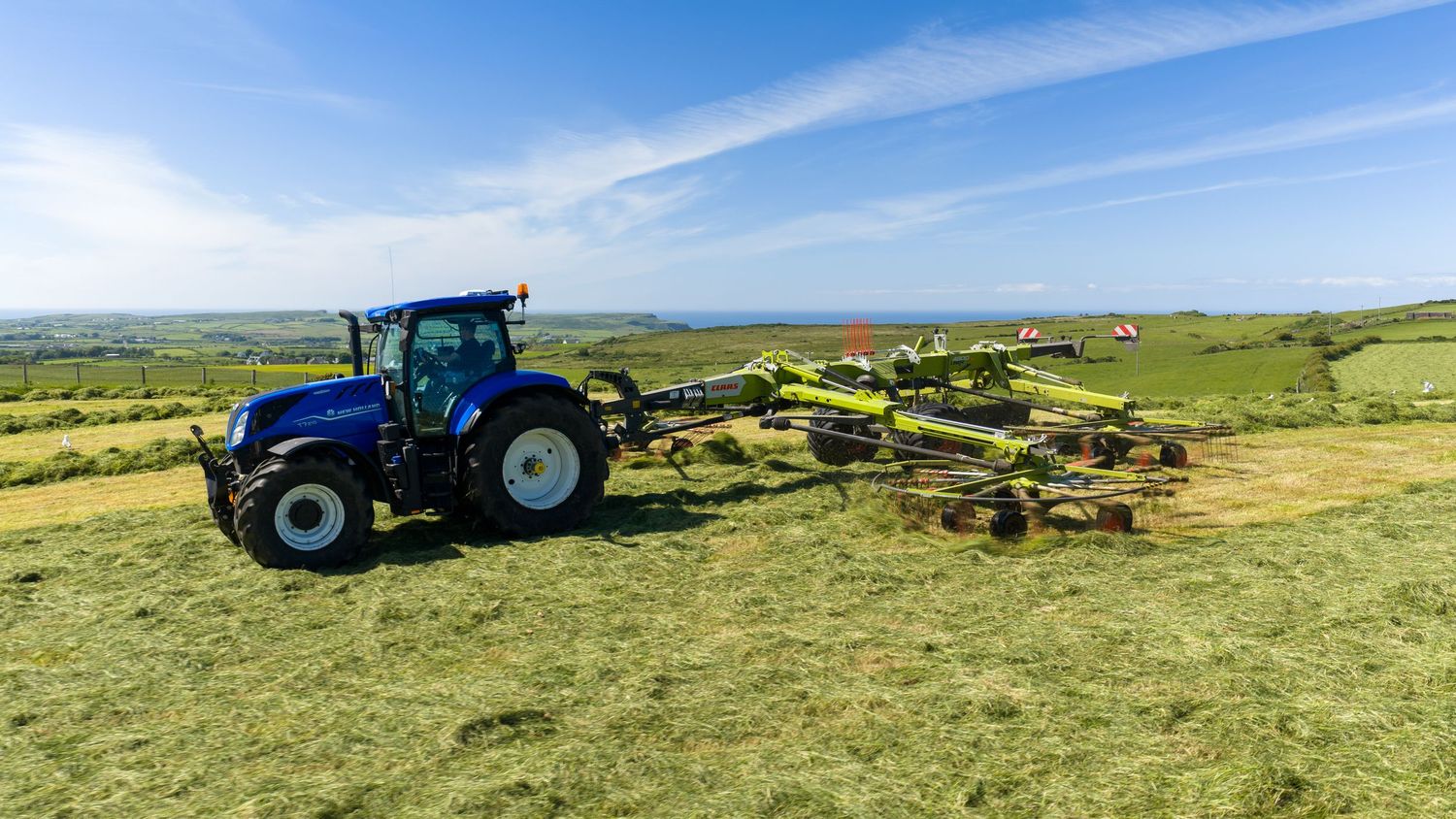 Blue New Holland tractor pulling a green hay rake across a lush green field under a clear blue sky.