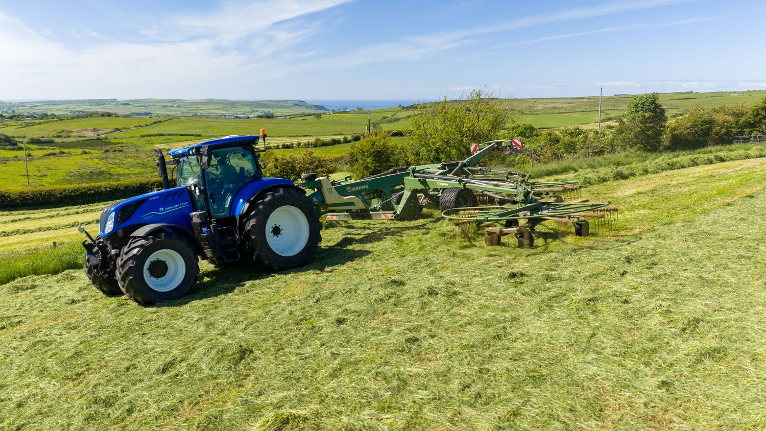 Blue New Holland tractor pulling green hay rake across lush green field with scenic countryside in background.