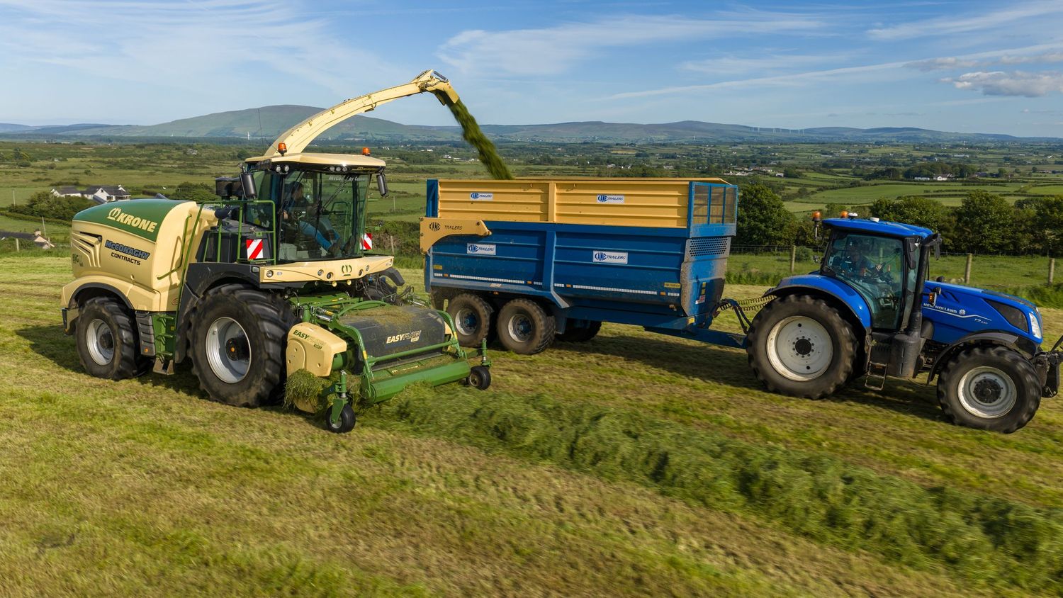 Green forage harvester transferring silage into blue tractor trailer in lush Irish countryside farmland.