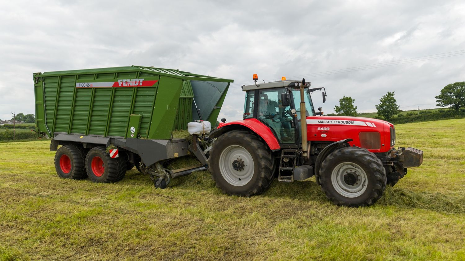 Red Massey Ferguson tractor pulling a green Fendt trailer on a farm field during hay harvesting season.