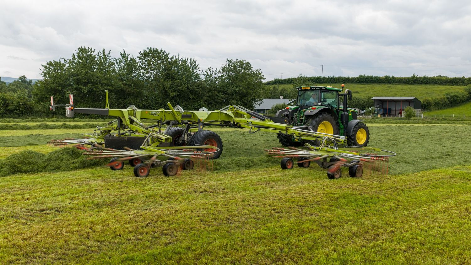 Green tractor with CLAAS hay rakes working in a lush field, gathering cut grass on an overcast day.