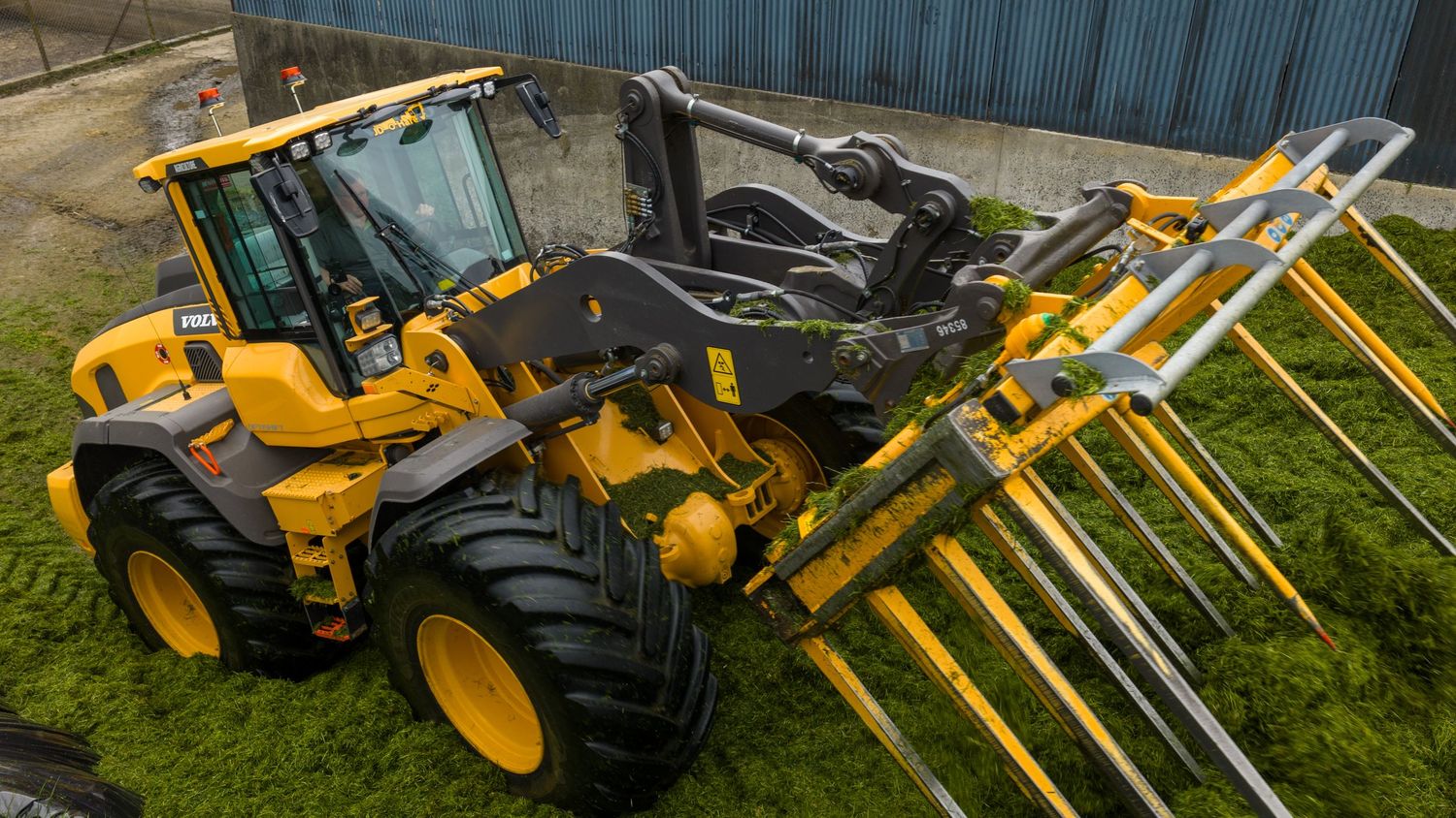 Yellow wheel loader with green hay fork attachment parked on grass near a building.