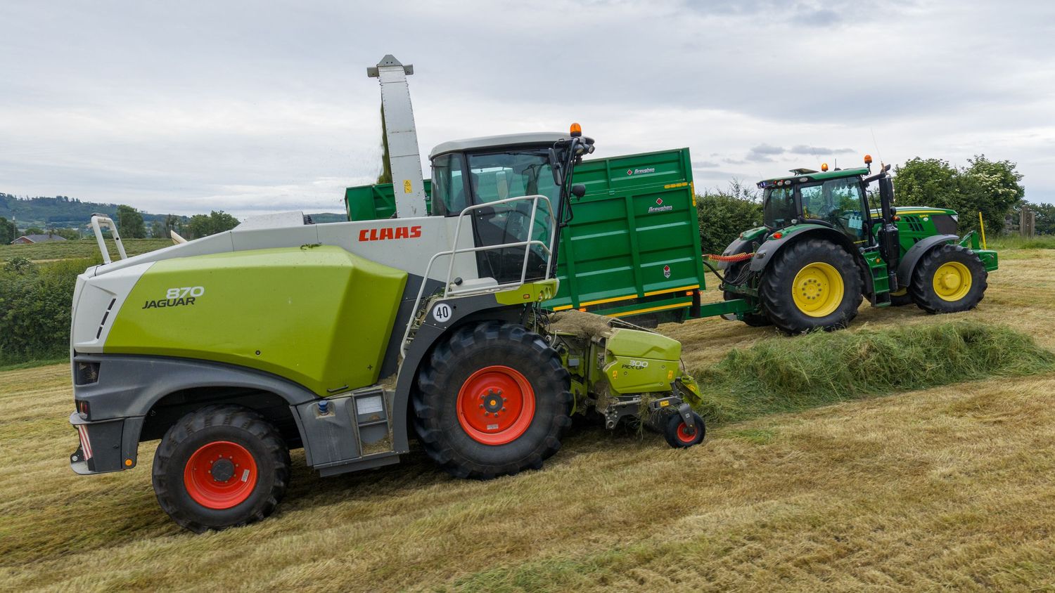 CLAAS harvester and John Deere tractor with green trailer collecting hay in a lush farm field.