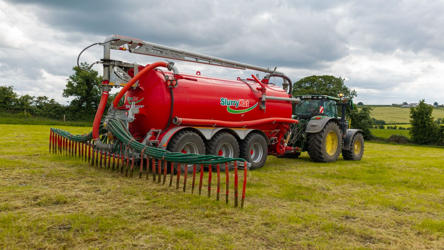 Red agricultural slurry tanker with trailing hose applicator attached to a green tractor in a field.