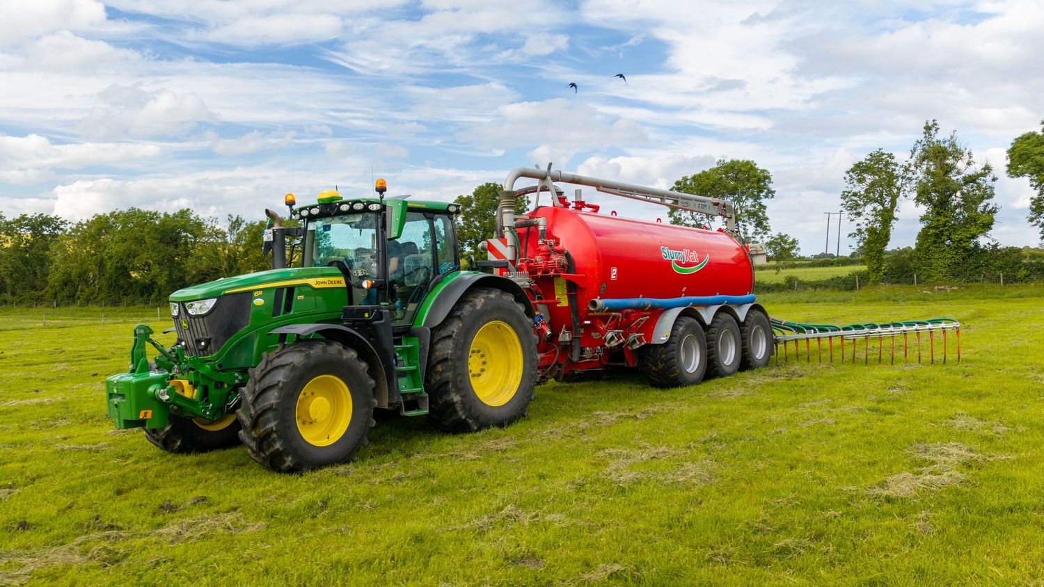 Green John Deere tractor pulling a red slurry tanker on a lush green farm field under a cloudy sky.