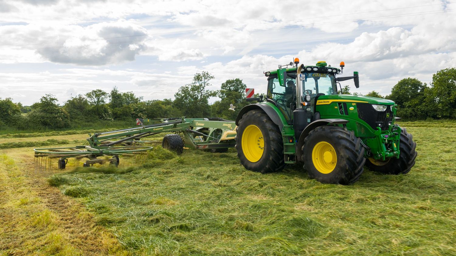 Green John Deere tractor with hay rake attachment working in a field, collecting cut grass on a cloudy day.
