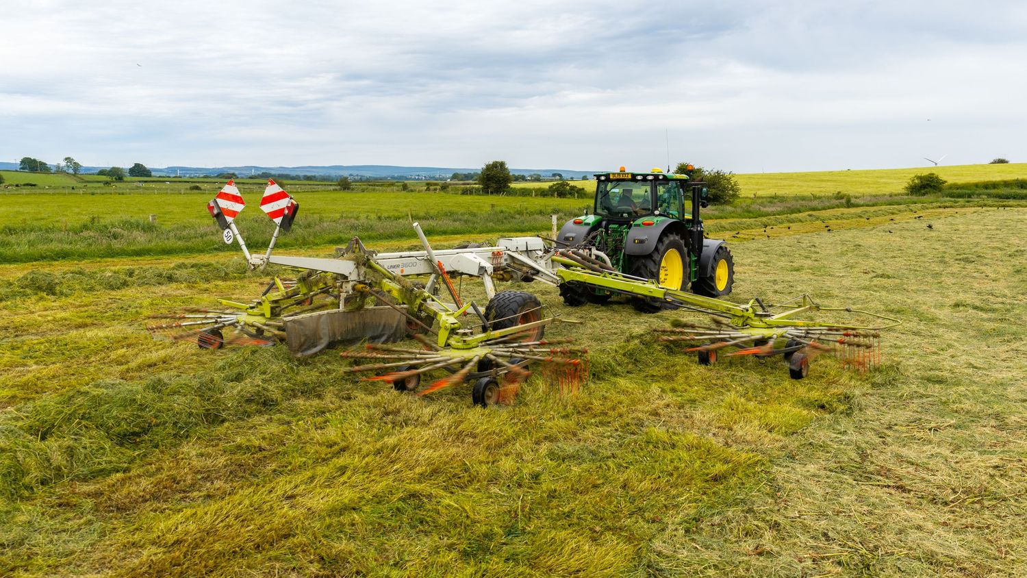 Green tractor pulling a Claas hay rake machine across a lush agricultural field under cloudy skies.