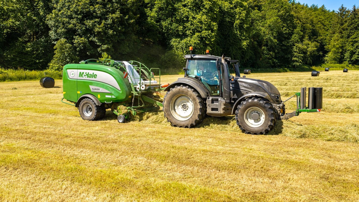 A black Valtra tractor with a green McHale round baler harvesting hay in a sunny field.
