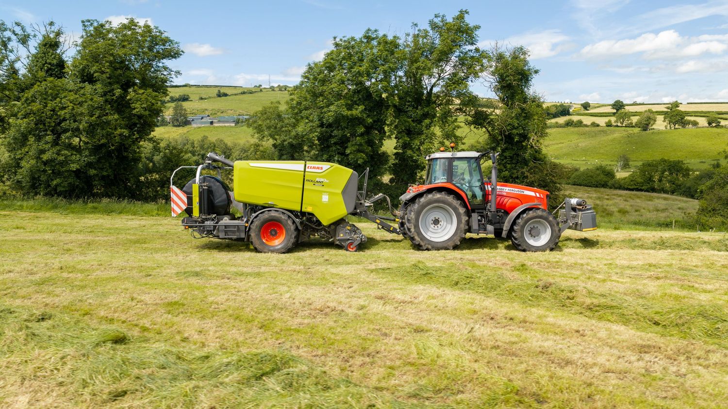 Red tractor pulling a green Claas round baler in a lush green hay field on a sunny summer day.
