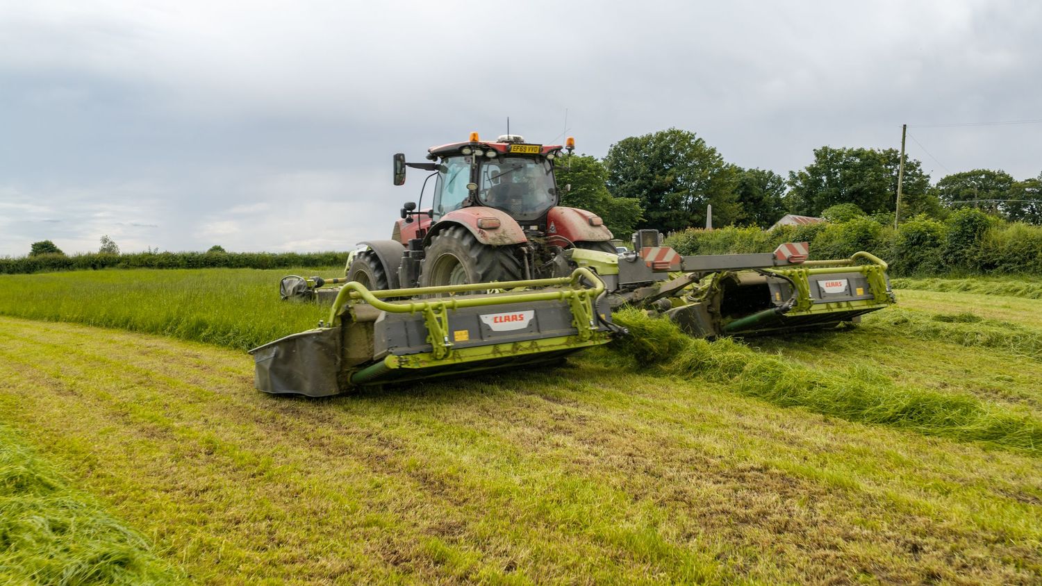 Red tractor with green Krone mower cutting grass in a lush farm field under overcast skies.