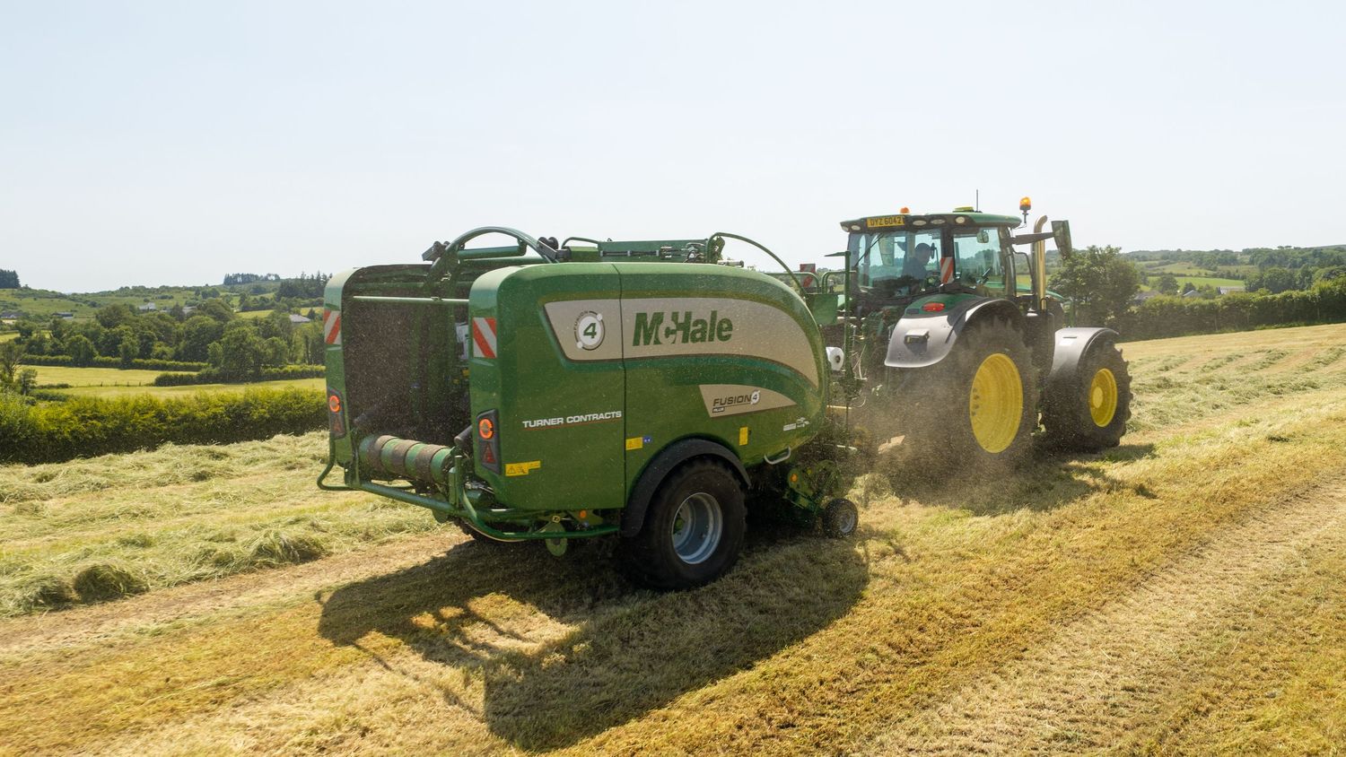 A green McHale baler attached to a tractor harvesting hay in a sunny field with lush green hedgerows in background.
