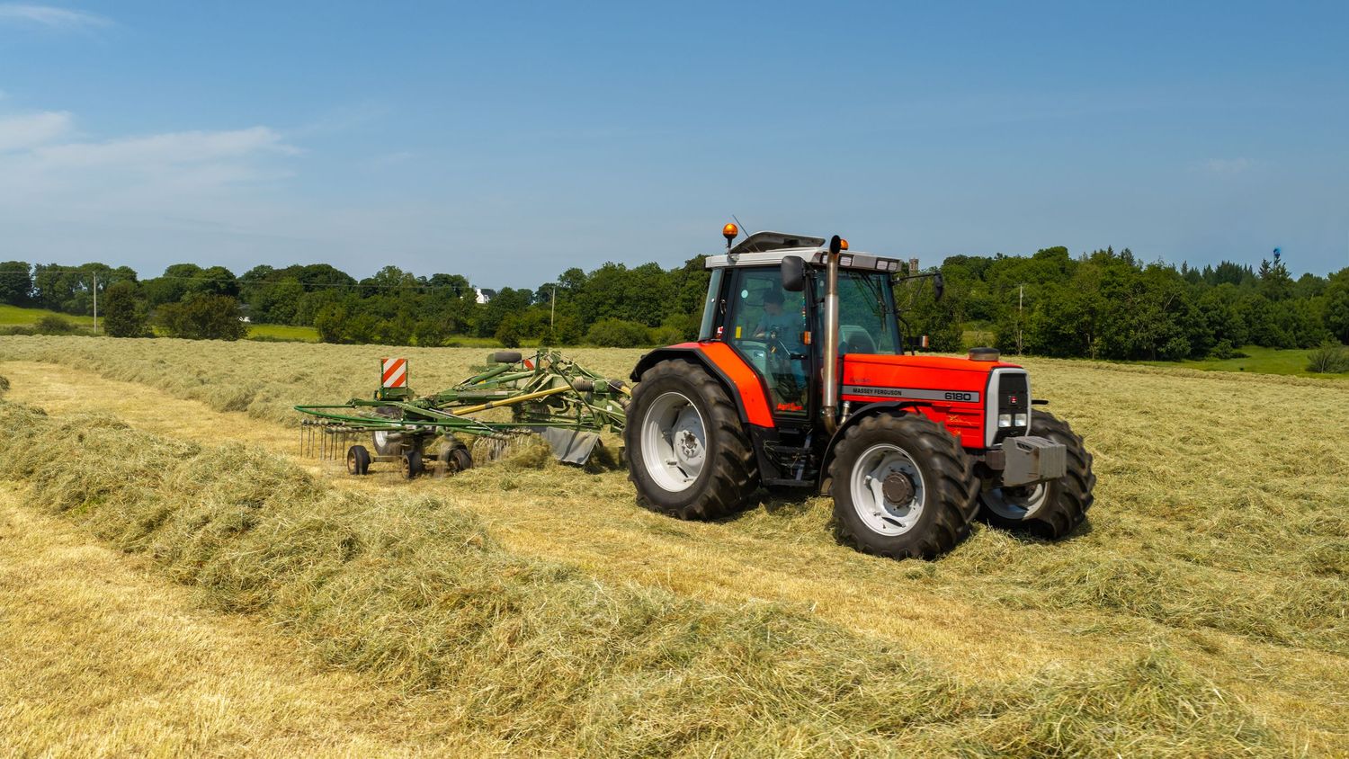 Red Massey Ferguson tractor raking cut hay in a sunny green field during summer harvest season.