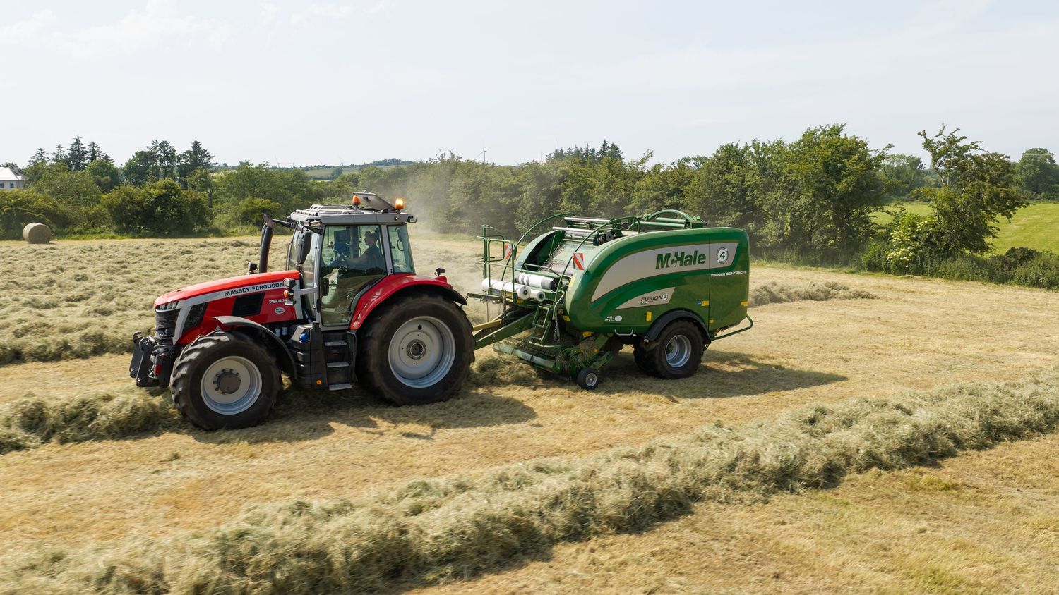 Red tractor pulling a green Vicon baler in a sunny hay field, collecting cut grass for silage baling.