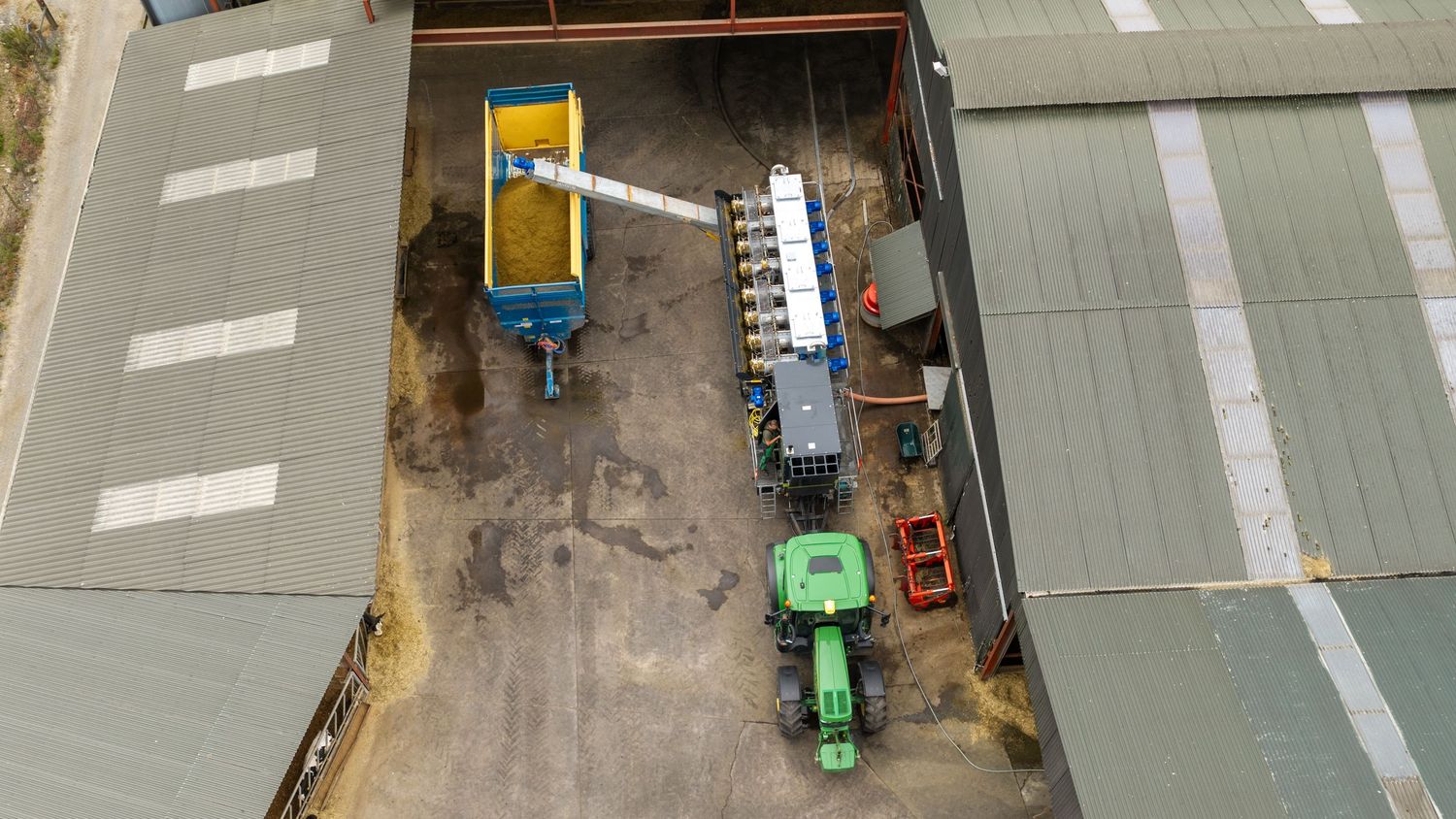 Aerial view of an industrial facility with green tractors, conveyor belt, and grain storage equipment on concrete.