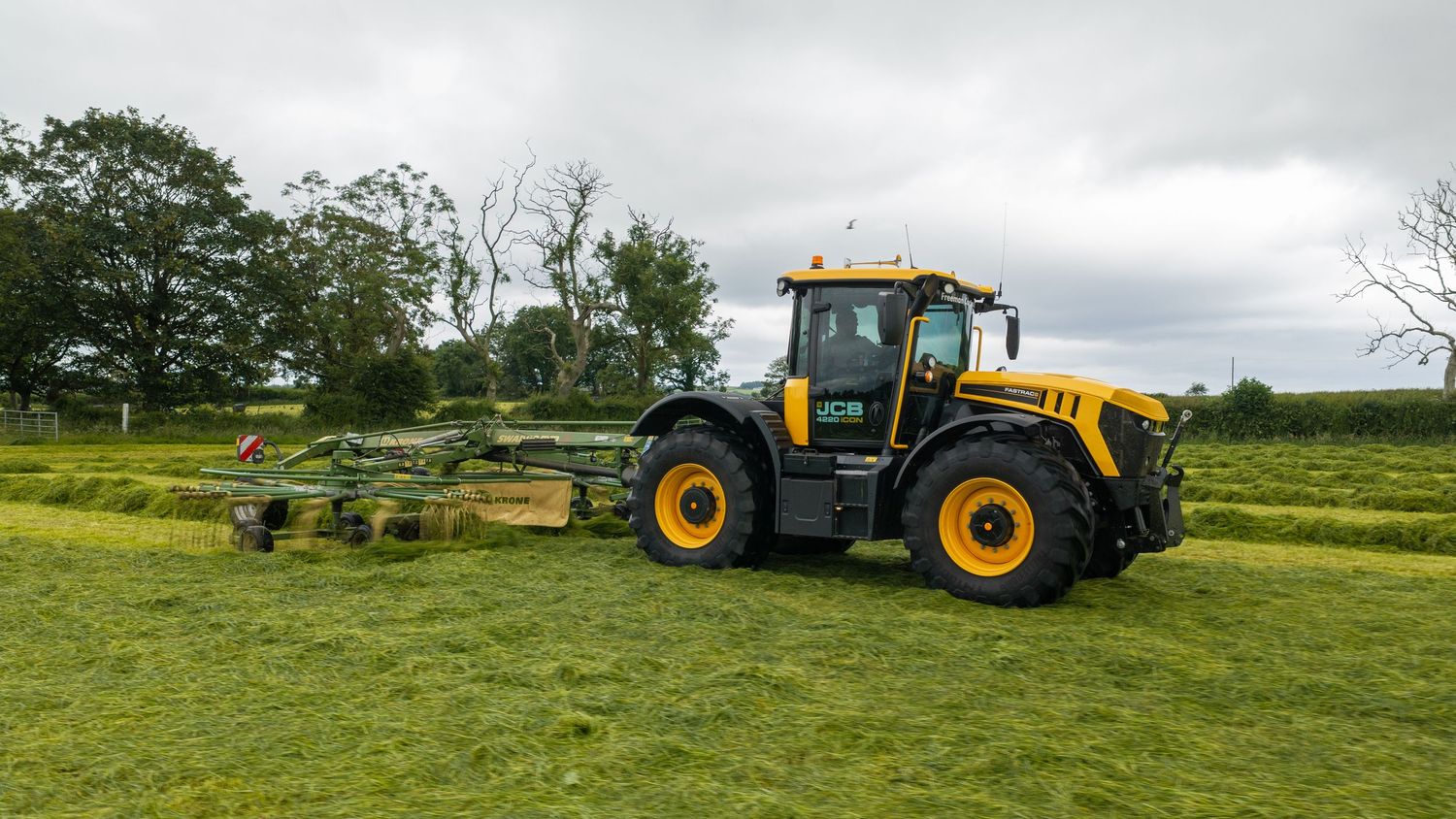 Yellow JCB tractor with mower attachment cutting green grass in a lush farm field on a cloudy day.