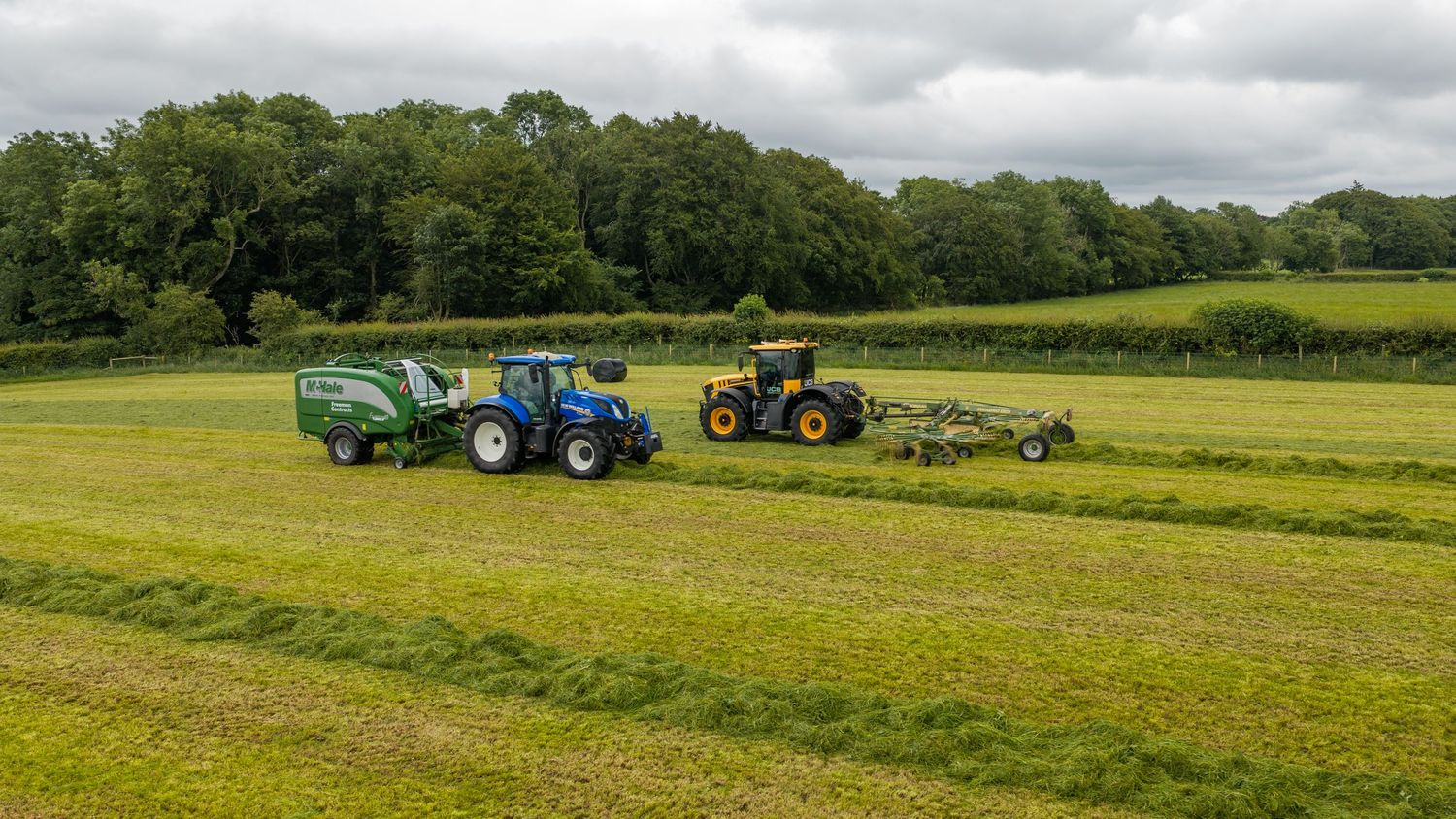 Two tractors working a green field, one baling hay, surrounded by lush trees under overcast skies.