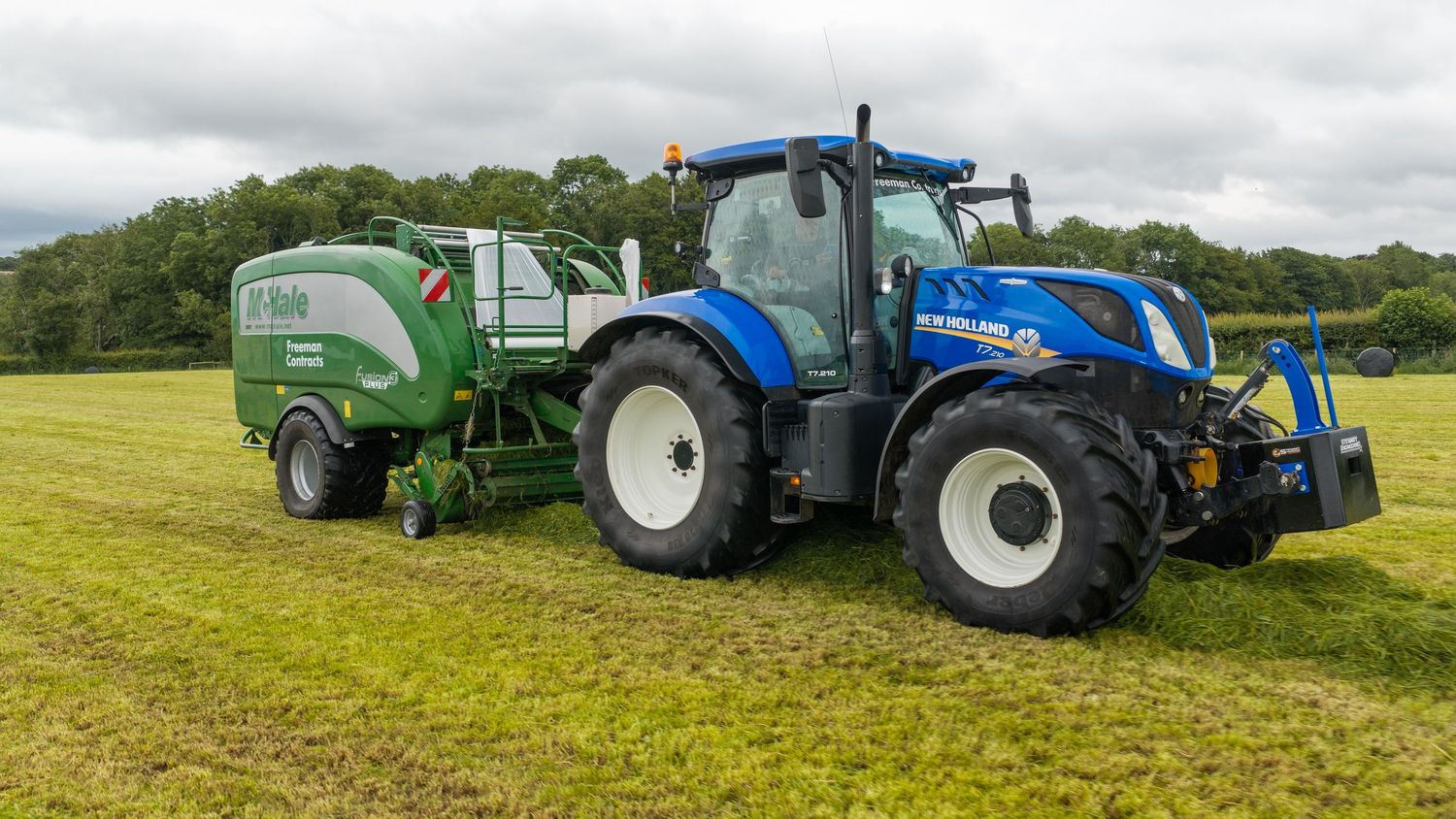 Blue New Holland tractor pulling a green McHale baler in a freshly cut green field.