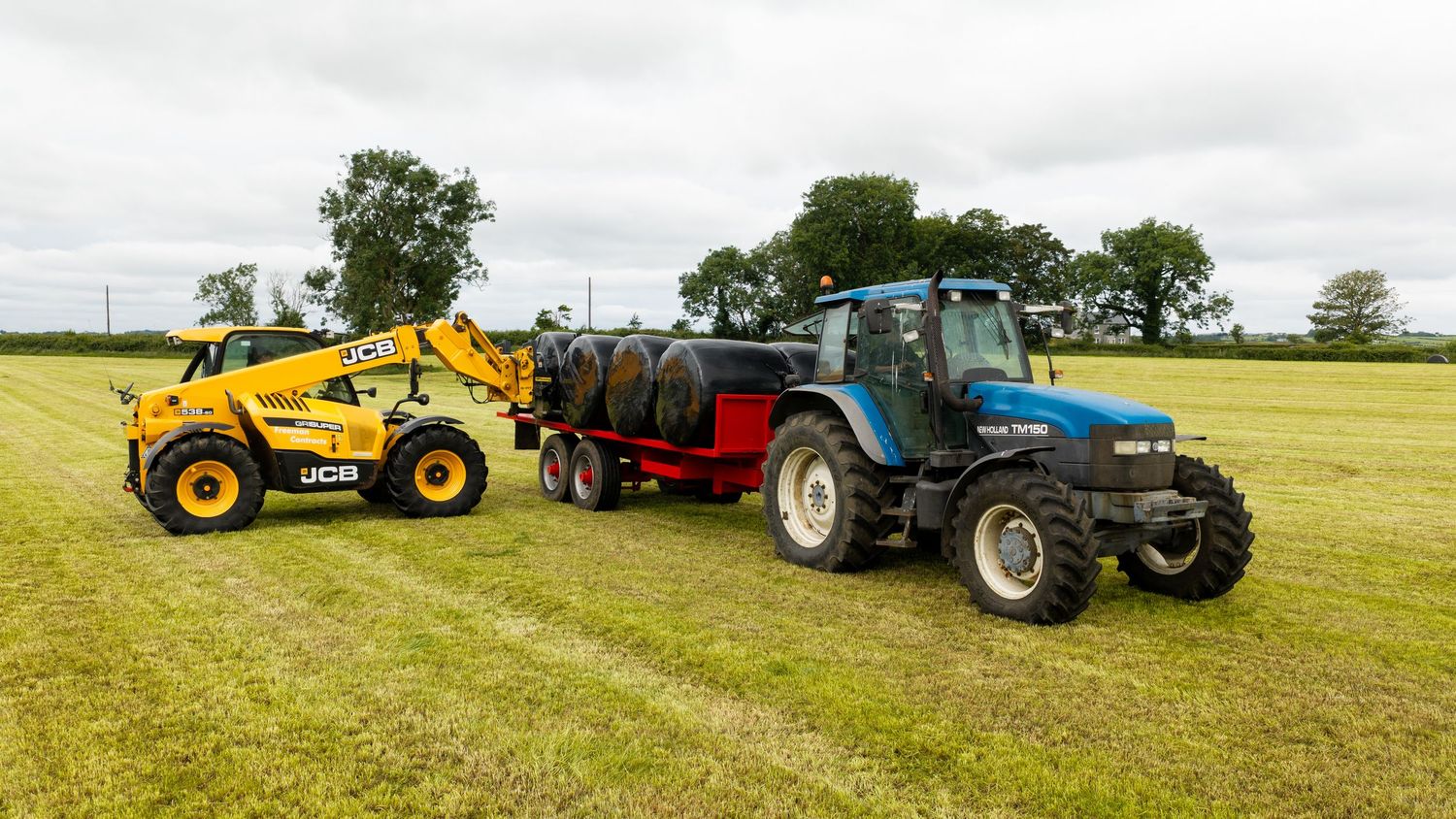 Blue New Holland tractor with red trailer and yellow JCB telehandler working in a green agricultural field.