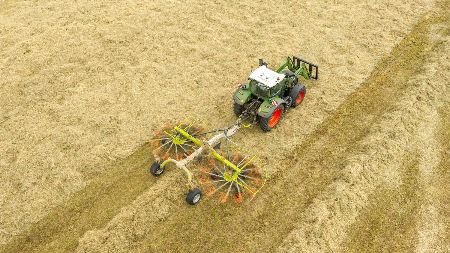 Aerial view of a green Fendt tractor with twin rotary rake attachment harvesting dry hay in a golden field.