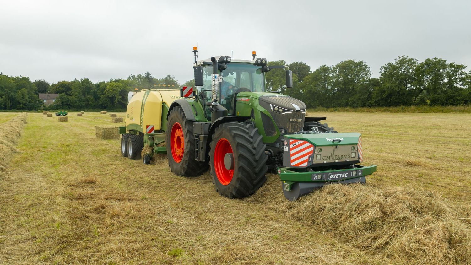 Green Fendt tractor with red wheels pulling a yellow baler on a harvested field with hay bales and trees in background.