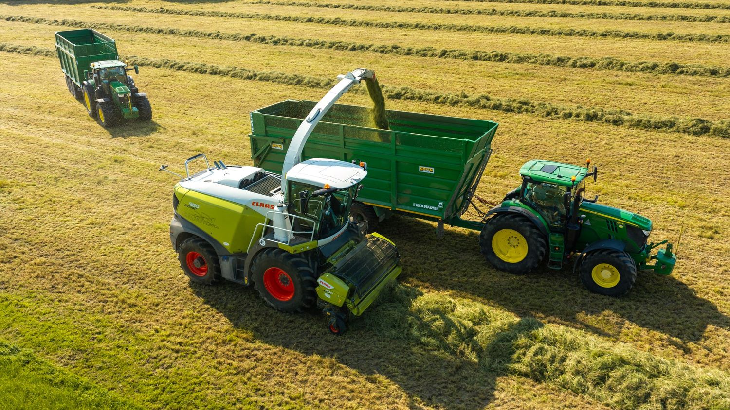 Aerial view of green Claas and John Deere tractors harvesting crops in a golden field during sunny conditions.