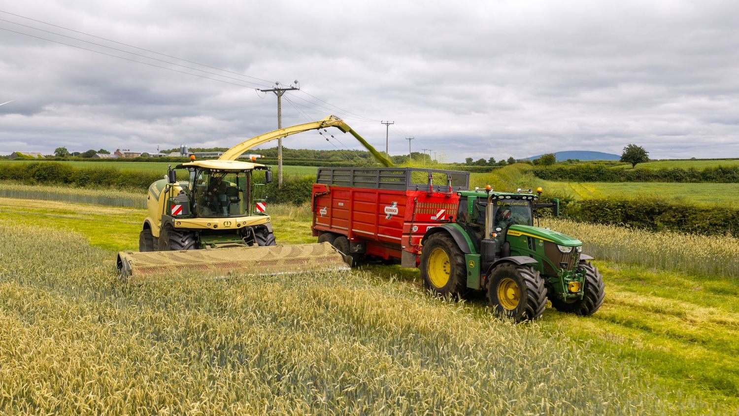 Green John Deere tractor with red trailer harvesting crops in a lush field using a yellow forage harvester.
