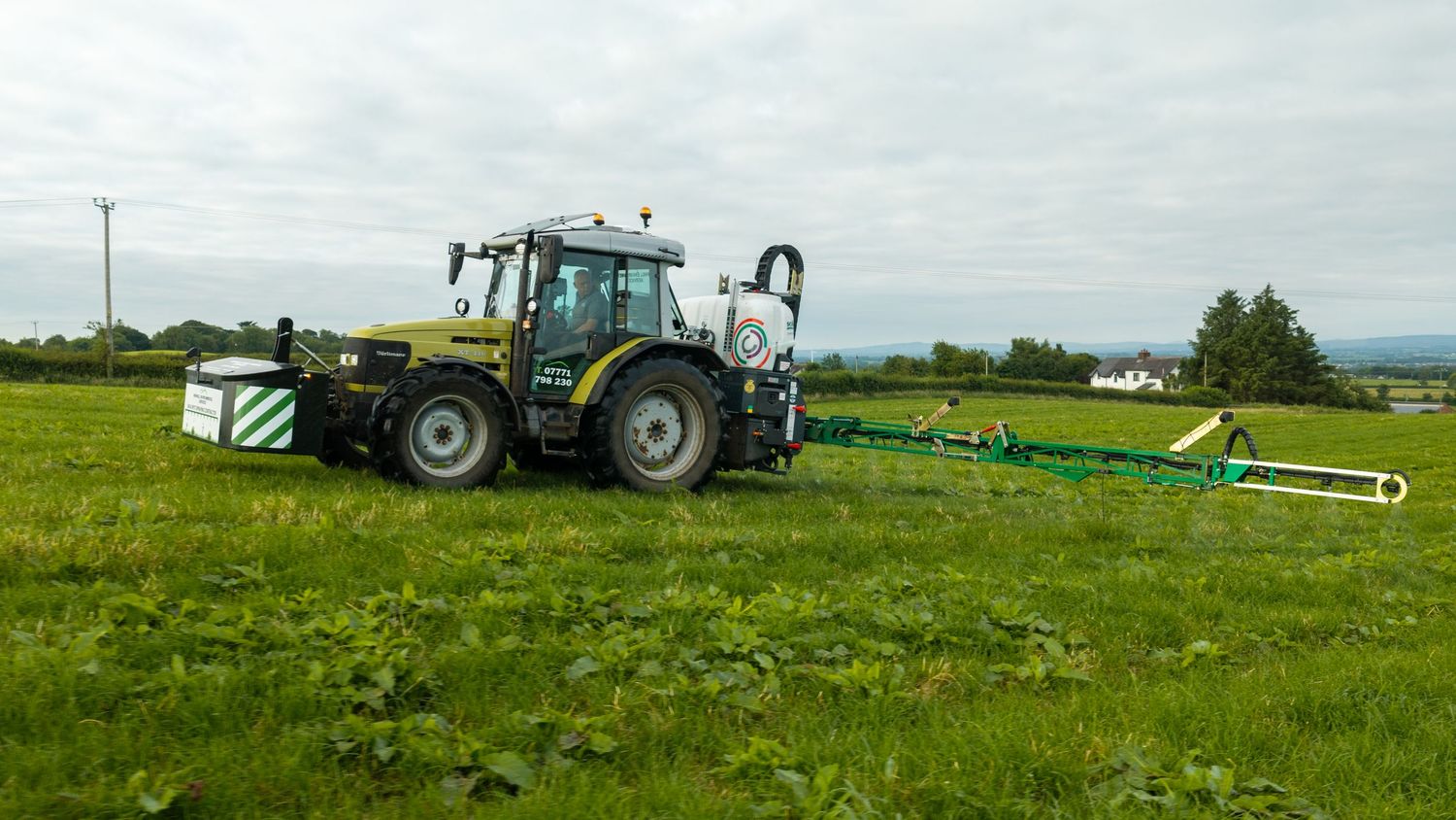 Green tractor with boom sprayer applying pesticides on lush farmland under overcast skies.
