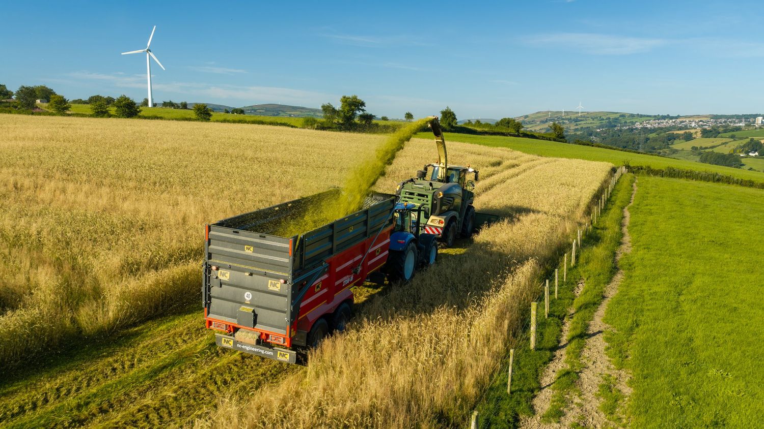 Aerial view of a green tractor harvesting crops in a lush field with a wind turbine in the background.