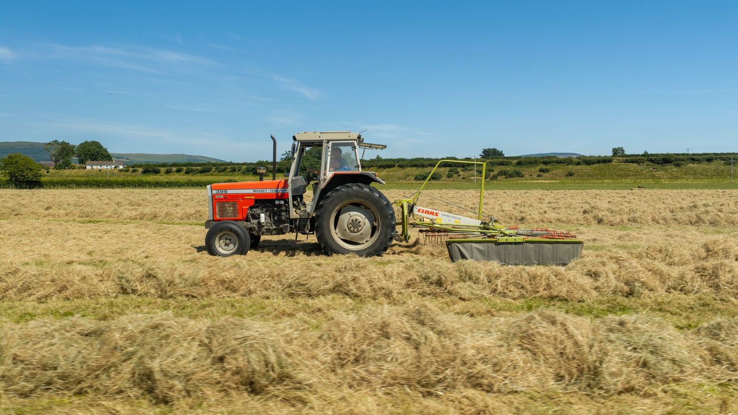 Red Massey Ferguson tractor haymaking in a sunny rural field with blue skies and green countryside in background.