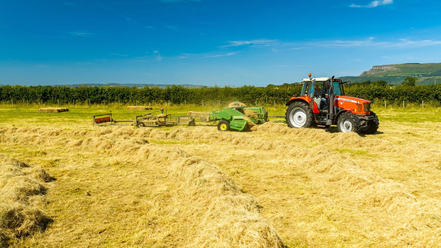 Red tractor baling hay in a sunny green field with rolling hills and trees in the background.