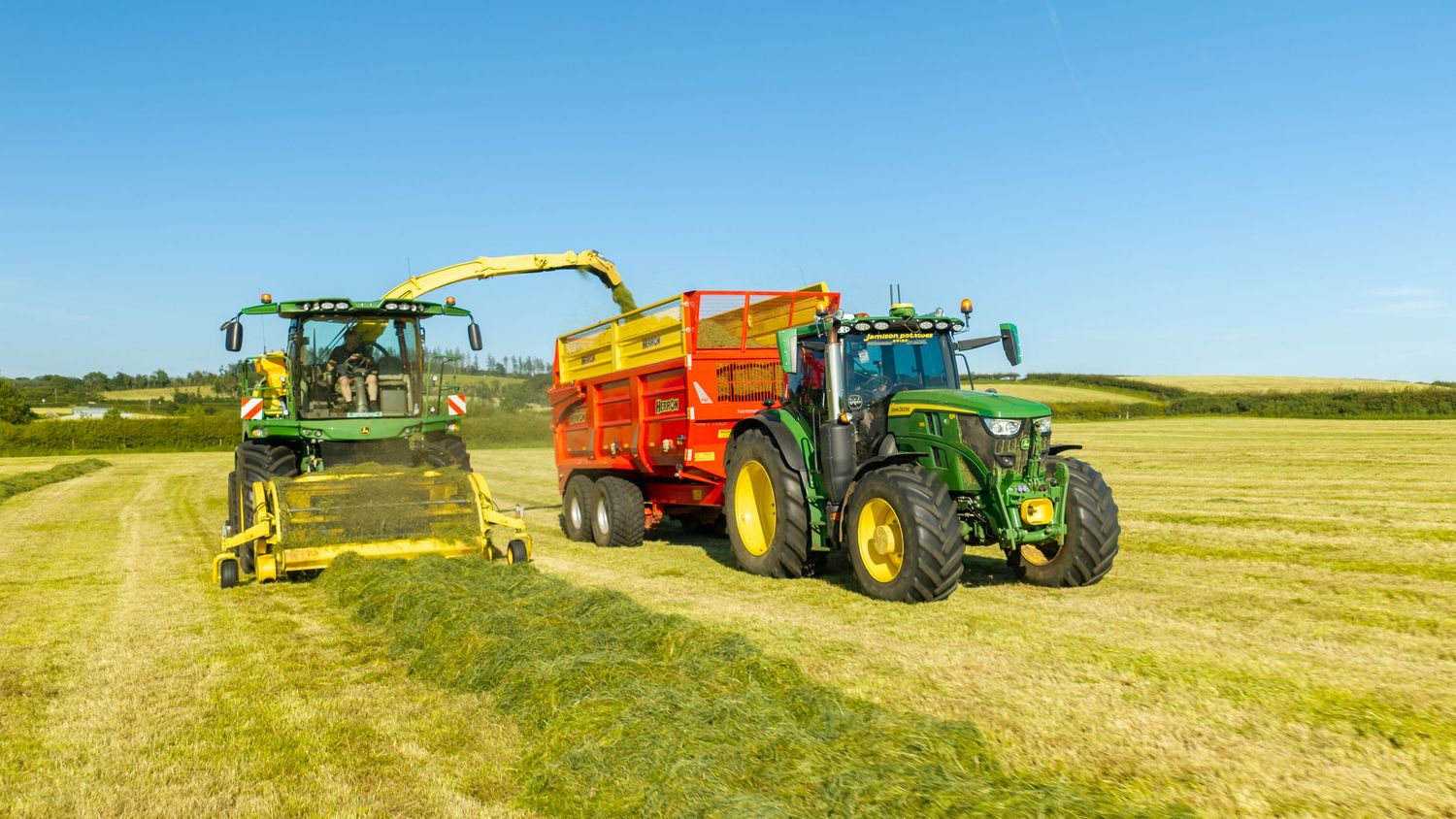 Two John Deere tractors harvesting silage in a sunny green field, with a forage harvester and orange trailer.