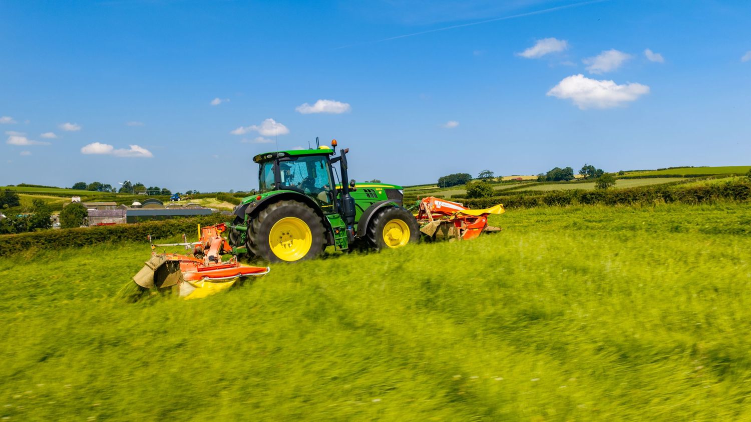 Green John Deere tractor mowing lush green fields on a sunny day in the countryside.