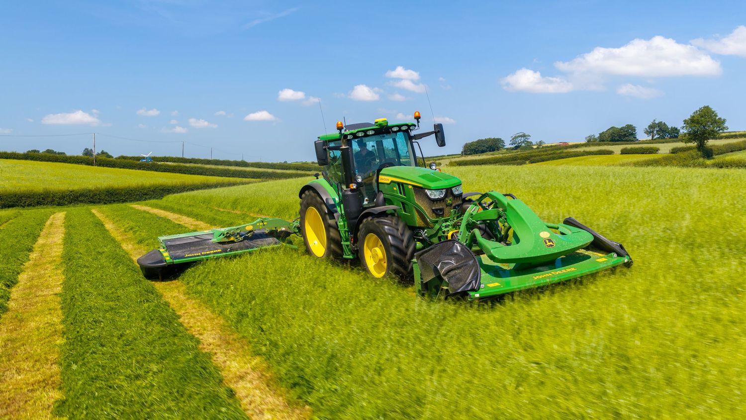 John Deere green tractor mowing a lush grass field on a sunny day with blue skies and rolling countryside.