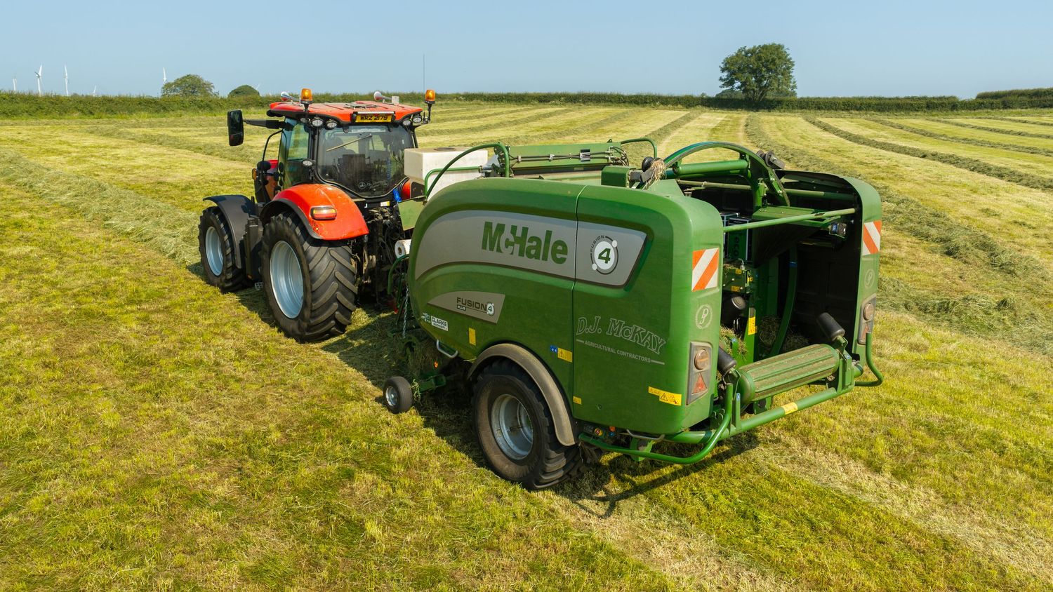 M-Hale round baler attached to a red tractor harvesting hay in a green field on a sunny day.