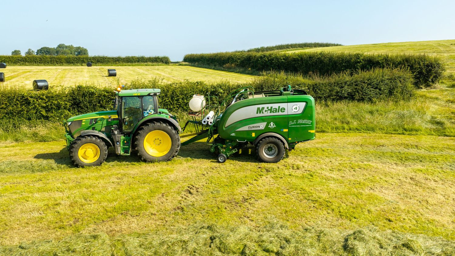 John Deere tractor pulling a McHale baler in a green field during hay baling season on a sunny day.