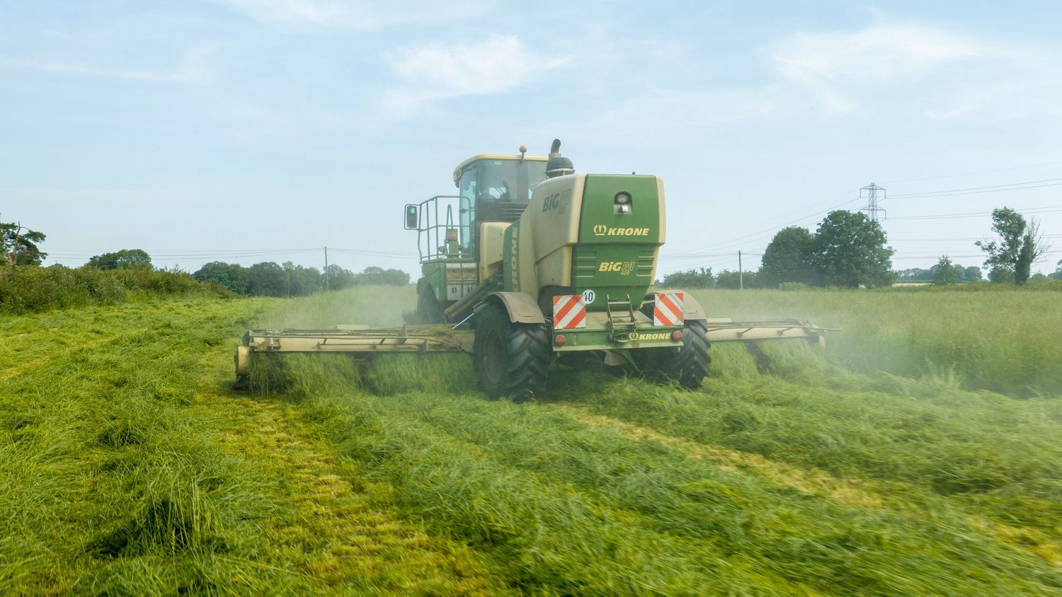 Green combine harvester cutting grass in a lush field on a sunny day with trees in the background.