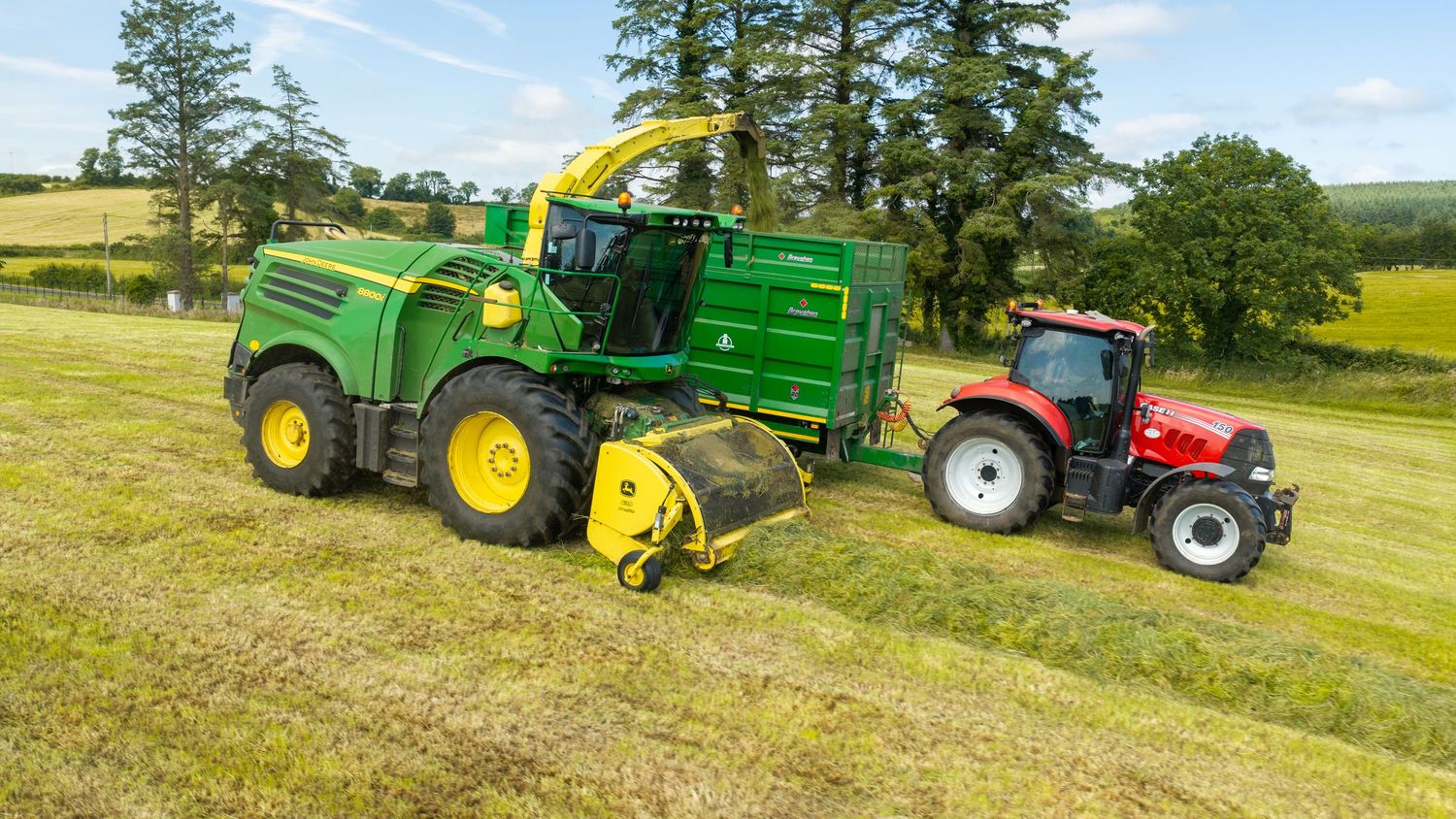 Green John Deere forage harvester working alongside a red tractor in a sunny field with trees in the background.