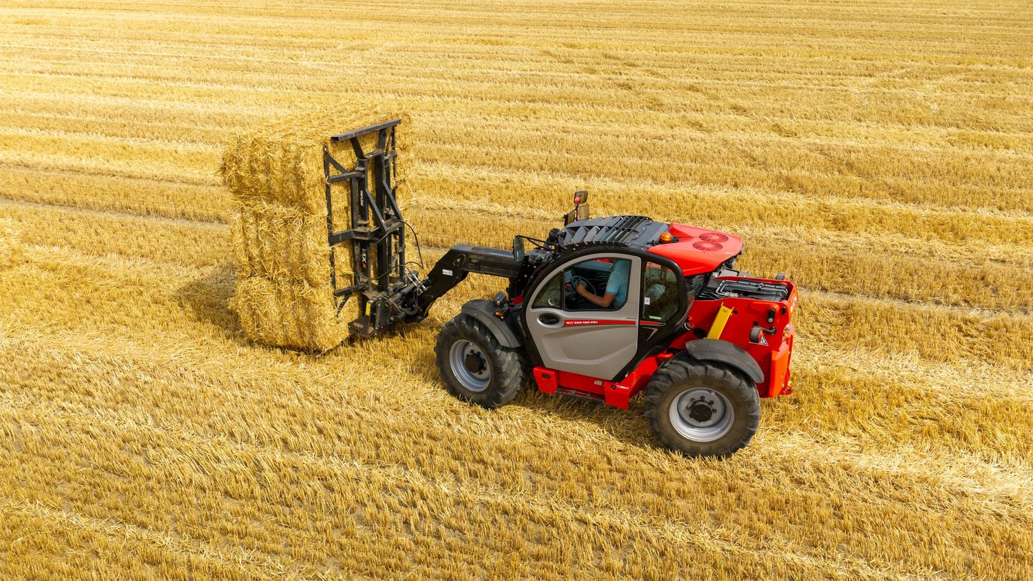 Red telehandler lifting a large hay bale in a golden wheat field, aerial view of agricultural harvesting equipment.